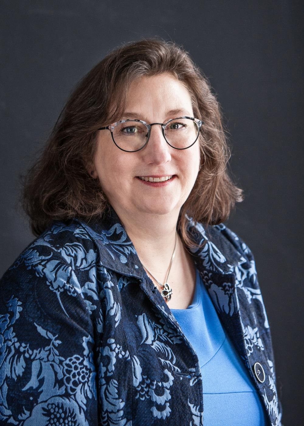 Judge Elect Jessica Skemp is wearing a dark blue suit coat with light blue flowers throughout the coat and a light blue shirt underneath. She is sitting in front of a dark background smiling. 