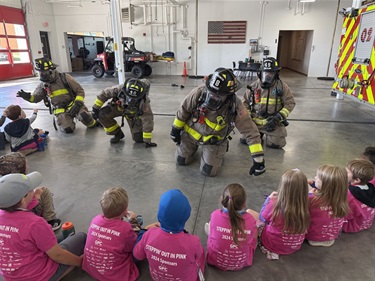 four firefighters in full gear crawl to a group of visiting preschoolers to give high fives.