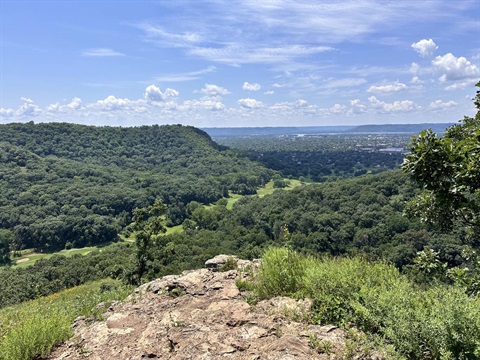 A scenic view from a rocky overlook of a lush green valley with a distant cityscape under a blue sky with scattered clouds.