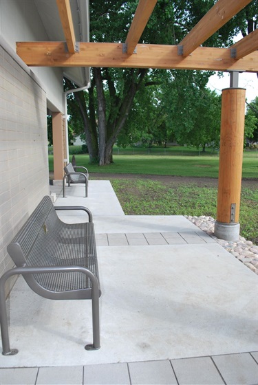 Two empty metal benches sit on a concrete patio under a wooden pergola with a large tree in the background.