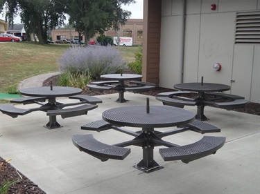 Four dark metal picnic tables with attached benches are arranged on a concrete patio next to a building and a garden of lavender flowers.