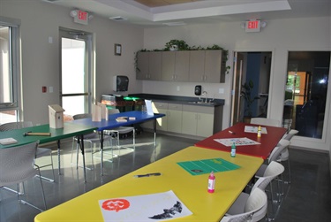 A brightly lit room with multiple tables set up for crafts, including a yellow table with art supplies and paper, a red table with craft supplies, and a green table with a birdhouse kit and wooden blocks. A kitchenette area with cabinets and a sink is visible in the background.