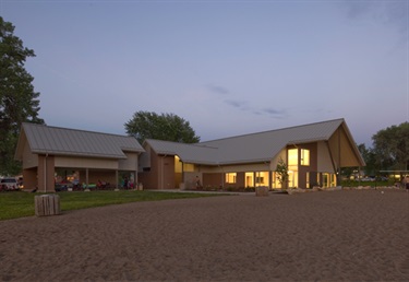 A modern building with a metal roof and large windows is lit up at dusk, with covered picnic areas and trees in the background, and a sandy foreground.
