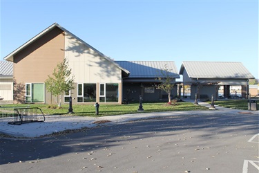 A modern building with a distinctive pitched roof, large windows, and a covered outdoor seating area. A bicycle rack is visible in the foreground, and the ground is scattered with fallen leaves, suggesting an autumn setting.