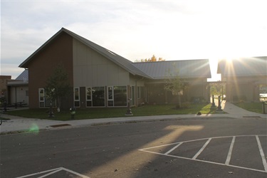 A modern building with a metal roof and large windows, bathed in the warm glow of the setting sun. The building features a combination of brown and light-colored siding, with trees and grass in the foreground. A paved parking lot with white markings is visible in front of the building.