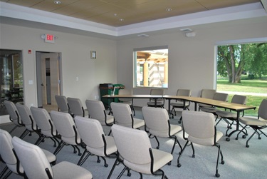 A meeting room with rows of beige chairs facing a light blue wall. Large windows on the left look out onto a grassy area with a large tree.