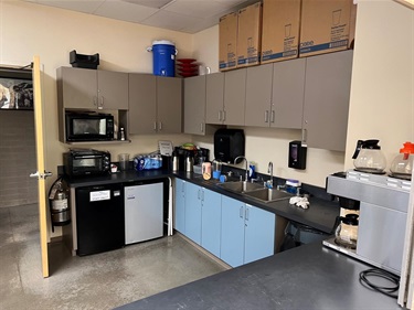 A breakroom kitchen area with a sink, microwave, two mini-fridges, coffee makers, and cabinets stacked with boxes.