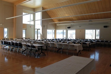 An event hall is set up with many tables covered in white tablecloths and chairs arranged around them, ready for guests. Sunlight streams in through large windows, illuminating the polished wooden floor.