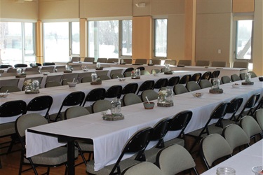 An event hall is set up with many tables covered in white tablecloths and chairs arranged around them, ready for guests. Sunlight streams in through large windows, illuminating the polished wooden floor.