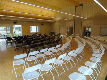 Rows of empty white and black folding chairs are set up in a large room with a wooden floor and a wood-paneled ceiling with modern lighting.