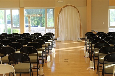 An indoor wedding ceremony setup with rows of chairs facing a draped archway. Wooden letter blocks spelling 'GARRETT' and 'BRUCE' are visible in the window.