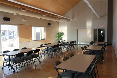 A view of a large room with multiple tables and chairs set up, suggesting a meeting or event space. The room has a high, sloped ceiling with exposed wooden beams and ductwork. Large windows on the left let in natural light, reflecting off the polish.