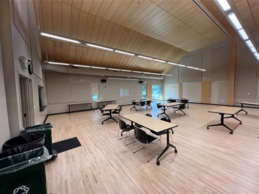 A spacious room with light wood floors and a vaulted ceiling with wood paneling. The room is furnished with multiple tables and chairs, set up for a meeting or event.