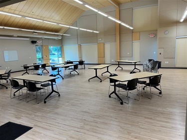 A large, empty room with light wood floors, featuring multiple tables and chairs set up for an event. Some tables are arranged in rows, while others are grouped. A stack of chairs is visible in the background. The room has a high, sloped ceiling with modern lighting and large windows letting in natural light.