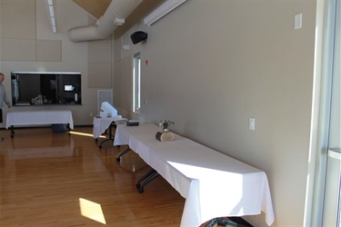 Long tables covered with white tablecloths are set up in a room with a wooden floor and beige walls. A small floral arrangement sits on one table. A counter with a window in the background.
