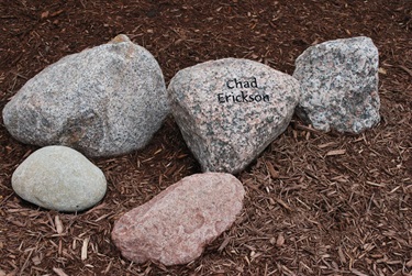 A stone engraved with the name Chad Erickson is surrounded by other natural stones on a bed of brown mulch.
