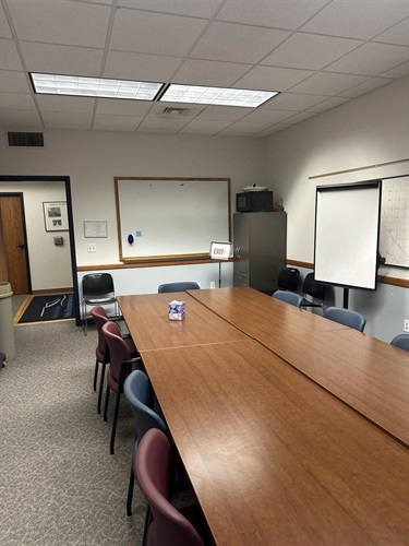 A long wooden conference table is surrounded by chairs. A whiteboard is on the wall, and a projector screen is set up in the corner.