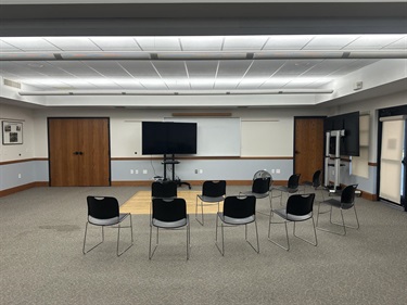 A conference room with a large screen monitor on a stand, a whiteboard, and several chairs arranged in a circle on a carpeted floor.