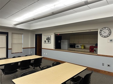 A view of a meeting room with tables and chairs, and a serving counter with a refrigerator, microwaves, and cabinets visible beyond.