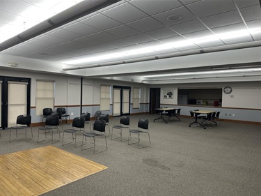 A large room with rows of black chairs facing a wooden stage area, and tables with chairs set up in the back near a serving counter.