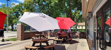 An outdoor seating area with picnic tables under white and red umbrellas. People are seated at one table, and another person stands nearby. The area is paved, with brick walls and green trees in the background.