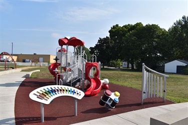 A colorful playground with a large red and white climbing structure, a rainbow-colored keyboard, and a drum set on a rubber surface with a grassy area and trees in the background.