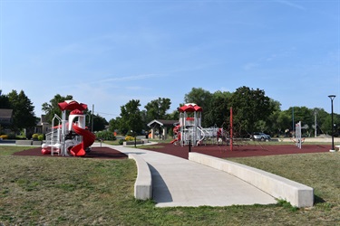 A modern playground with red and white structures, slides, and climbing equipment on a clear day with a concrete pathway leading through the grassy area.