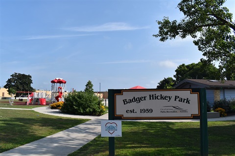 A sign for Badger Hickey Park, established in 1959, with a playground and building in the background.