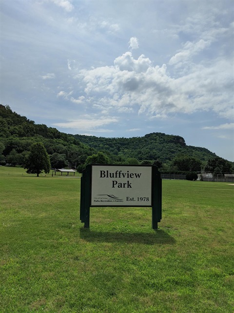 Sign for Bluffview Park, established in 1978, with a grassy field and tree-covered hills in the background under a cloudy sky.