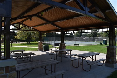 A covered picnic area with multiple picnic tables and benches, overlooking a tennis court and grassy area.