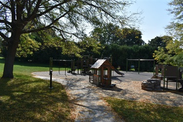 A wooden playground with swings, a slide, and a small playhouse is set in a park with trees and a paved path.