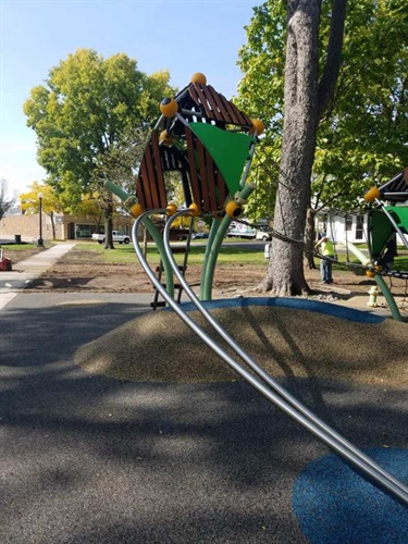 A modern playground structure with a wooden treehouse-like design and green accents, set in a park with trees and a paved area.