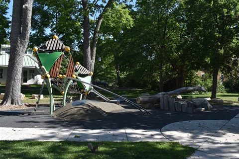 A modern playground with green and brown structures, slides, and natural wood elements, set among trees on a sunny day. A squirrel is visible on the grass in the foreground.