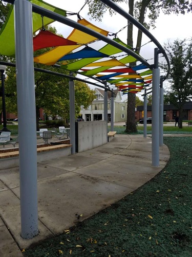 A curved walkway passes under a canopy of colorful shades supported by gray poles. The walkway is bordered by grass and a concrete wall with benches.