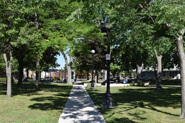 A paved walkway leads through a park with mature trees and green grass. In the background, buildings and people are visible.