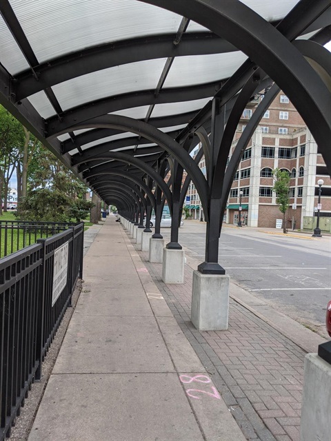 A covered walkway with a black arched frame and a translucent roof, extending down a sidewalk next to a road and a building.