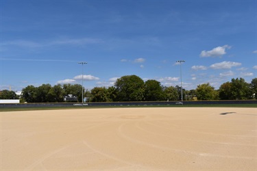 A view of an empty baseball field with a sand infield and green outfield, under a bright blue sky with scattered clouds. Lights and trees line the background, with a scoreboard reading 