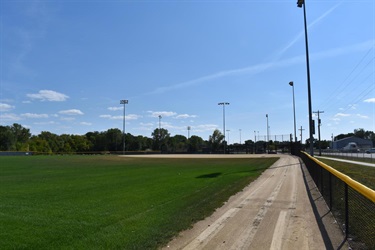 A baseball field with green grass, a dirt infield, and stadium lights under a blue sky with scattered clouds.