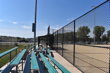 Empty bleachers with teal seats line a baseball field with a chain-link fence under a clear blue sky.