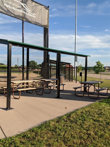 Covered picnic tables and benches are set up next to a chain-link fence under a clear blue sky.