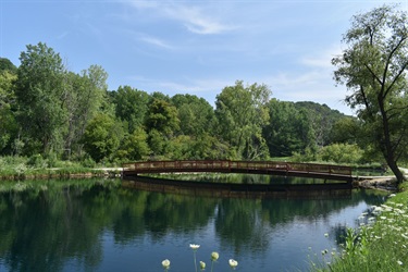 A wooden arched bridge spans a calm body of water, reflecting the lush green trees and blue sky on the opposite bank.