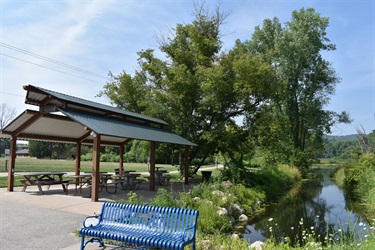 A blue metal bench sits on a paved path next to a covered picnic area with picnic tables and a small stream with lush green trees in the background.
