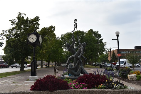 A bronze sculpture of figures holding lacrosse sticks reaching upwards, set against a backdrop of trees, a street clock, and buildings.