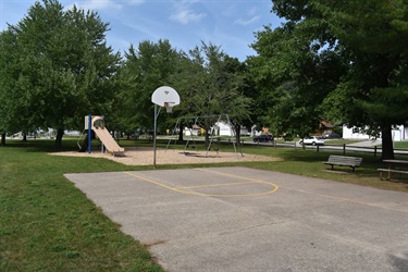 A park scene featuring a basketball court, a slide, swings, and picnic tables surrounded by trees and grass.