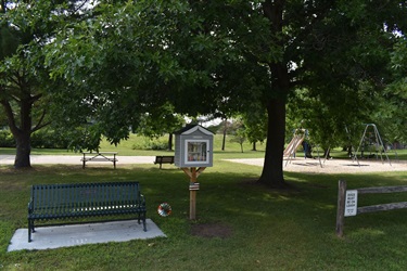 A small library box on a wooden post is situated in a park next to a green bench and a playground with swings and slides.