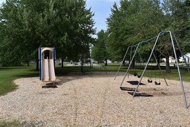 A playground with a slide and swings on a wood chip surface, surrounded by green trees and grass.