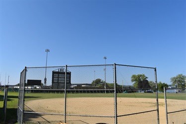 A baseball field with a chain-link fence in the foreground and a scoreboard in the background.