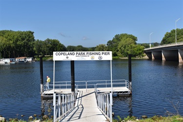 A fishing pier at Copeland Park extends over a dark blue body of water, with a person standing on the pier. A large bridge spans the water in the background, and trees line the distant shore.