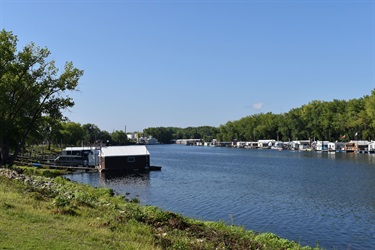 A wide, calm river with many houseboats docked on the left and right sides, surrounded by trees under a clear blue sky.