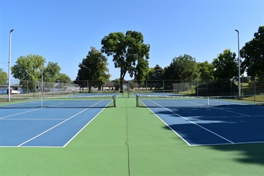 A wide view of multiple blue and green tennis courts with nets, surrounded by trees and under a clear blue sky.